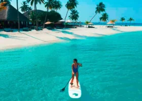 Una donna in costume pratica stand up paddle (SUP) su una tavola bianca nella laguna turchese cristallina, con la spiaggia bianca, le palme da cocco e gli edifici dal tetto in strame del resort sullo sfondo. Constance Halaveli, Halaveli, Atollo di Ari Nord, Maldive.