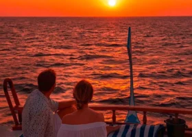 Una coppia abbracciata su cuscini a righe blu e bianche osserva il sole che cala sull'Oceano Indiano tingendo il cielo di arancio e rosso durante una crociera al tramonto. Constance Halaveli, Isola di Halaveli, Atollo di Ari Nord, Maldive.