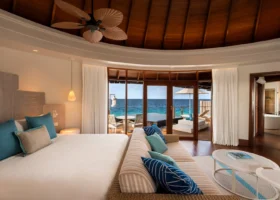 Interno di una water villa dall'alto soffitto in legno con ventilatore a foglie di palma, letto king con cuscini turchesi e blu, divano e vetrate scorrevoli aperte sul deck con piscina privata affacciato direttamente sull'oceano. Constance Halaveli, Halaveli, Atollo di Ari Nord, Oceano Indiano.