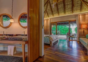 Interni luminosi di una Beach Villa del Constance Moofushi con alto soffitto in makuti e travi in legno, letto king size con copriletto a fantasia tropicale, pavimento in legno scuro lucido e ventilatore a pale; sulla sinistra la sala da bagno con doppi lavabi in pietra su mobile in legno e due specchi tondi con cornice intrecciata; ampie porte scorrevoli aprono sulla vegetazione e sulla spiaggia dell'isola di Moofushi, Atollo di Ari Sud, Maldive.
