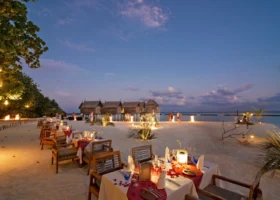 Allestimento per una cena romantica a lume di candela sulla spiaggia di sabbia bianca del Constance Moofushi al crepuscolo, con tavoli vestiti di bianco e runner bordeaux, petali di rose, calici in cristallo e lanterne accese; sullo sfondo le strutture overwater dal tetto sagomato e il cielo blu del tramonto sull'oceano, a Moofushi, Atollo di Ari Sud, Maldive.