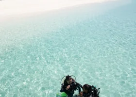 Due subacquei in muta con bombole e pinne gialle durante un corso sub nelle acque cristalline e poco profonde della laguna turchese del Constance Moofushi; alle spalle la spiaggia di sabbia bianca con lettini, un lungo pontile e le ville overwater, a Moofushi, Atollo di Ari Sud, Maldive.