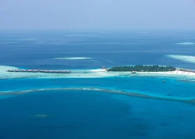Panorama aereo dell'isola corallina del Constance Moofushi ripresa da lontano: distesa di palme sul lato destro, lunga fila di ville overwater collegate da un pontile sul reef, spiaggia di sabbia bianca e ampie fasce di laguna turchese alternate a canali blu profondo; Moofushi, Atollo di Ari Sud, Maldive, Oceano Indiano.