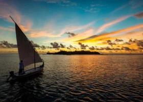 Dhoni tradizionale maldiviano con grande vela bianca e un uomo in piedi sulla prua che naviga al tramonto sulle acque calme che riflettono la luce dorata; all'orizzonte la silhouette dell'isola con le palme sotto un cielo acceso di arancio, oro e rosa, al Constance Moofushi, Atollo di Ari Sud, Maldive.