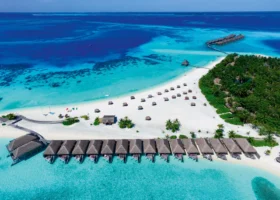 Veduta aerea della spiaggia del Constance Moofushi con una lunga lingua di sabbia bianca, fila di ville overwater dai tetti in makuti su pontile, ombrelloni intrecciati sulla sabbia, fitta vegetazione di palme e area per gli sport acquatici; laguna turchese e barriera corallina blu intenso a Moofushi, Atollo di Ari Sud, Maldive, Oceano Indiano.