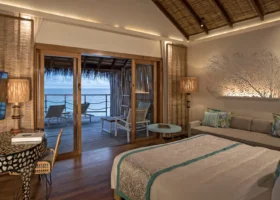 Interni eleganti di una Water Villa overwater del Constance Moofushi con letto king size, soffitto a travi in legno e makuti, illuminazione soffusa, lampade da terra in rattan intrecciato, decorazione a parete a ventaglio di corallo retroilluminata e scrittoio tondo con intarsi in madreperla; le porte scorrevoli in vetro aprono sulla terrazza privata con lettini e vista diretta sull'oceano al crepuscolo, a Moofushi, Atollo di Ari Sud, Maldive.