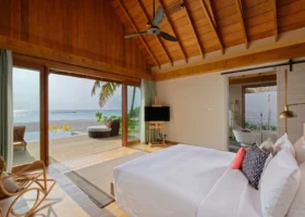 Interno della beach villa con alto soffitto a volta in legno e ventilatore, letto king con biancheria bianca e cuscini corallo e blu; ampie vetrate scorrevoli aperte sul deck in legno con daybed in rattan e piscina privata a sfioro, accesso diretto alla spiaggia e al mare, e sulla destra il bagno open-air con vasca freestanding. Faarufushi, Atollo di Raa, Maldive.