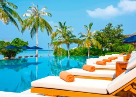Piscina a sfioro con lettini in legno dai materassi bianchi, teli arrotolati arancioni e ombrelloni blu lungo il bordo; alte palme da cocco e vegetazione tropicale si specchiano nell'acqua, mentre oltre la piscina si aprono la laguna e il cielo azzurro. Emerald Faarufushi, Atollo di Raa, Maldive.