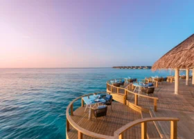 Terrazza del ristorante overwater con deck curvilineo in legno, tavoli apparecchiati e sedie intrecciate affacciati sull'oceano nella luce pastello del tramonto; il tetto in strame sulla destra e, all'orizzonte, la fila di ville overwater sospese sulla laguna. Faarufushi, Atollo di Raa, Maldive.