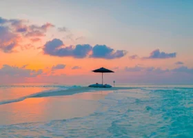 Angolo romantico su una lingua di sabbia bianca al tramonto: un ombrellone e morbidi pouf allestiti sulla riva mentre il cielo si tinge di arancio, rosa e viola e le onde leggere lambiscono la battigia turchese. Emerald Faarufushi, Atollo di Raa, Maldive.