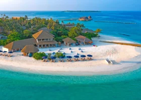 Veduta aerea della spiaggia di sabbia bianca con gli edifici dal tetto in strame del beach club, ombrelloni blu e lettini allineati, un catamarano in secca e un fitto palmeto; la laguna turchese sfuma nel blu dell'oceano con le strutture overwater sullo sfondo. Faarufushi, Atollo di Raa.