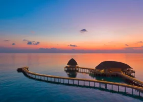 Veduta al crepuscolo delle strutture overwater dal tetto in strame collegate da un pontile in legno illuminato che si snoda sulla laguna calma, mentre il cielo pastello arancio e viola si riflette sull'acqua immobile. Emerald Faarufushi, Atollo di Raa, Maldive.
