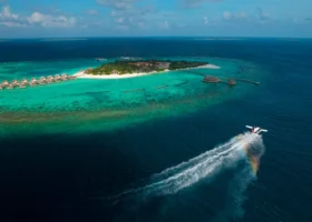 Veduta aerea dell'isola di Faarufushi con la fila di ville overwater dal tetto in strame, la barriera corallina affiorante e la spiaggia bianca circondata dalla laguna turchese; in primo piano un idrovolante decolla lasciando una scia spumeggiante attraversata da un arcobaleno. Atollo di Raa, Maldive, Oceano Indiano.