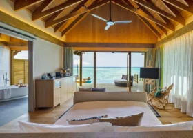 Interno della water villa overwater con soffitto a volta in legno chiaro e illuminazione calda, letto king e zona living aperti tramite vetrate scorrevoli sul deck sospeso con daybed e ombrellone affacciato sulla laguna turchese; a sinistra il bagno con vasca freestanding e il mobile bar. Faarufushi, Atollo di Raa, Oceano Indiano.