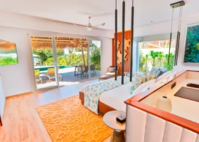 Interno luminoso di una beach villa dal design contemporaneo, con letto king, tessili turchesi a motivi marini, tappeto arancione e opere d'arte a tema corallo; le vetrate scorrevoli si aprono sul deck con lettini e piscina privata affacciata sul verde. Emerald Maldives, Fasmendhoo, Atollo di Raa, Maldive.