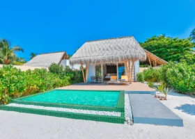 Esterno di una beach villa dal tetto in strame con piscina privata dal rivestimento a mosaico verde bordata di ciottoli bianchi, deck in legno con lettini gialli e area lounge, immersa tra la vegetazione tropicale e la sabbia bianca. Emerald Maldives Resort & Spa, Isola di Fasmendhoo, Atollo di Raa.