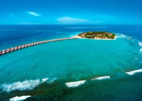 Veduta aerea dell'isola di Fasmendhoo con la lunga fila di water villa dal tetto rosso allineate sul pontile che si protende nella laguna turchese, la barriera corallina affiorante con le onde e il fitto verde tropicale dell'isola. Emerald Maldives, Atollo di Raa, Maldive.