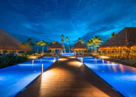 Veduta notturna all'ora blu delle piscine a sfioro illuminate ai lati di un camminamento in legno, con padiglioni e gazebo dal tetto in strame dalle calde luci dorate, palme da cocco e il cielo blu intenso del crepuscolo. Emerald Maldives Resort & Spa, Fasmendhoo, Atollo di Raa.