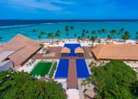 Veduta aerea dell'area centrale del resort con le grandi piscine dai rivestimenti blu, un campo in erba, gazebo e padiglioni dal tetto in strame, un fitto filare di palme e la spiaggia bianca che si affaccia sulla laguna turchese e sull'oceano. Emerald Maldives, Fasmendhoo, Atollo di Raa.