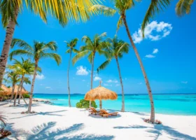 Spiaggia di sabbia bianca incorniciata da alte palme da cocco inclinate, con due lettini in legno e un ombrellone in paglia affacciati sulla laguna turchese cristallina sotto un cielo azzurro con nuvole leggere. Emerald Maldives, Isola di Fasmendhoo, Atollo di Raa, Maldive.