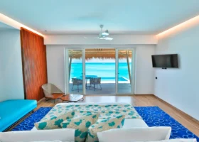 Interno di una water villa dal design contemporaneo con letto king, tessili turchesi a motivi marini e tappeto blu; le vetrate scorrevoli si aprono sul deck con piscina privata a sfioro e tavolo affacciati direttamente sulla laguna turchese. Emerald Maldives, Fasmendhoo, Atollo di Raa, Oceano Indiano.