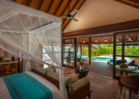 Interno di una beach villa con letto a baldacchino e zanzariera bianca, alto soffitto in legno con travi a vista e ventilatore, dettagli tessili turchesi; le vetrate scorrevoli si aprono sul deck con daybed e piscina privata immersa nel verde tropicale. Four Seasons Kuda Huraa, Atollo di Malé Nord, Maldive.