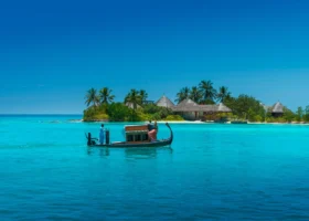 Tradizionale dhoni maldiviano dalla prua e poppa ricurve scivola sulla laguna turchese con a bordo una coppia e il barcaiolo in uniforme bianca; sullo sfondo l'isola-resort con ville dal tetto in strame e palme da cocco. Four Seasons Kuda Huraa, Atollo di Malé Nord, Oceano Indiano.