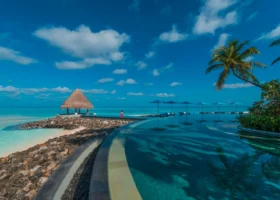 Lunga piscina a sfioro dal profilo curvilineo che costeggia una lingua di roccia con gazebo dal tetto in strame, ombrelloni blu e una donna in abito rosa che passeggia sul bordo; oltre, la laguna cristallina sopra il reef. Four Seasons Kuda Huraa, Atollo di Malé Nord, Maldive.