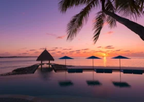 Tramonto rosa e arancio sulla piscina a sfioro che riflette il cielo, con gazebo dal tetto in strame in silhouette, ombrelloni blu e la fronda di una palma in primo piano mentre il sole cala sull'oceano. Four Seasons Kuda Huraa, Atollo di Malé Nord, Maldive.