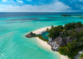 Veduta con drone della punta dell'isola-resort, con ville dal tetto in strame immerse nel palmeto, piscina a sfioro curvilinea e spiaggia bianca lambita da acque che sfumano dal turchese al blu profondo. Four Seasons Kuda Huraa, Atollo di Malé Nord, Maldive.