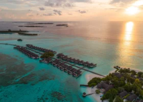 Veduta aerea al tramonto delle ville overwater dal tetto in strame disposte a spina di pesce lungo il pontile sulla laguna turchese, con il sole che si specchia sull'oceano dorato e l'isola coperta di palme. Four Seasons Kuda Huraa, Atollo di Malé Nord, Maldive.