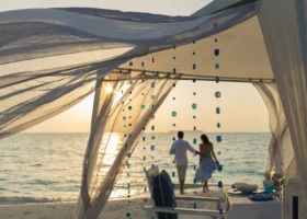 Allestimento romantico in spiaggia al tramonto sotto un gazebo di teli bianchi mossi dal vento e fili di perline blu e verdi; una coppia mano nella mano cammina verso il mare mentre il sole cala all'orizzonte e un tavolo apparecchiato attende sulla sabbia. Landaa Giraavaru, Atollo di Baa, Maldive.