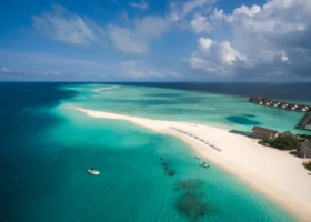 Veduta aerea di una lingua di sabbia bianca lambita da acque che sfumano dal turchese al blu profondo, con lettini allineati sulla riva, due piccole barche ormeggiate e le ville overwater del resort sulla destra. Four Seasons Landaa Giraavaru, Atollo di Baa, Maldive.