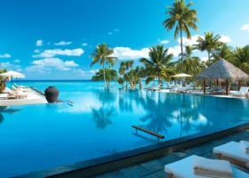 Piscina a sfioro che si fonde con l'oceano turchese all'orizzonte, con lettini e ombrelloni bianchi, palme da cocco e una sala dal tetto in strame; una grande giara in terracotta in primo piano. Four Seasons Landaa Giraavaru, Atollo di Baa, Riserva della Biosfera UNESCO, Maldive.