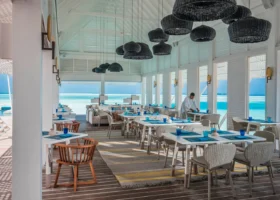 Interno del ristorante overwater Blu dalla struttura bianca e soffitto alto, con lampade a sospensione in fibra intrecciata nera e tavoli apparecchiati con bicchieri blu; un cameriere prepara i tavoli davanti alle ampie vetrate affacciate sulla laguna turchese. Landaa Giraavaru, Atollo di Baa, Maldive.