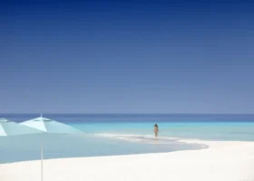 Spiaggia di sabbia finissima e bianchissima con una donna in bikini che cammina lungo la lingua di sabbia tra le acque turchesi, ombrelloni color acquamarina in primo piano e l'oceano blu all'orizzonte sotto un cielo terso. Four Seasons Landaa Giraavaru, Atollo di Baa, Maldive.