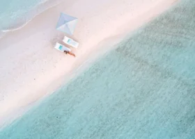 Veduta aerea dall'alto della battigia dove un ombrellone bianco e due lettini accolgono una persona in relax mentre un'altra fa snorkeling nell'acqua bassa e turchese, tra la sabbia bianca e i riflessi del reef. Four Seasons Landaa Giraavaru, Atollo di Baa, Maldive.