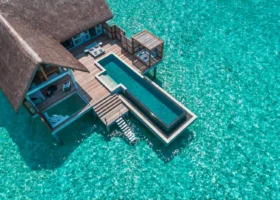 Veduta aerea dall'alto di una sunset water villa dal tetto in strame con lunga piscina privata a sfioro e ampio deck in legno, con la scala che scende direttamente nella laguna turchese cristallina dell'Oceano Indiano. Four Seasons Landaa Giraavaru, Atollo di Baa, Maldive.