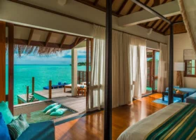 Interno di una water villa con letto a baldacchino e travi in legno, vetrate scorrevoli aperte sul deck sospeso con daybed e amaca affacciati sulla laguna turchese; dettagli tessili verde smeraldo e blu ne scaldano l'atmosfera. Landaa Giraavaru, Atollo di Baa, Maldive, Oceano Indiano.