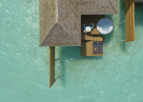 Veduta aerea dall'alto di una Aqua Villa overwater dal tetto in scandole di legno, con ampio deck che ospita jacuzzi rotondo, lettini e tavolo affacciati sulla laguna turchese cristallina attraversata dai pali di sostegno. Kandima Maldives, Isola di Kandima, Atollo di Dhaalu.