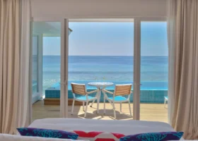 Vista dall'interno di una Aqua Villa attraverso le vetrate scorrevoli: tavolo e sedie sul deck in legno accanto alla piscina privata a sfioro dal mosaico turchese, affacciata direttamente sull'Oceano Indiano; in primo piano cuscini a motivi corallo. Kandima Maldives, Kandima, Atollo di Dhaalu.