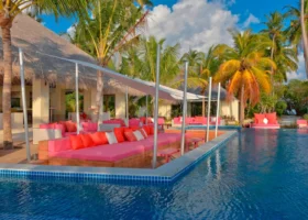 Angolo del beach club con divani e daybed dai cuscini fucsia e corallo affacciati sulla piscina a sfioro dal mosaico blu, sotto una tettoia in strame e tese bianche, tra alte palme da cocco e un cielo azzurro con nuvole. Kandima Maldives, Isola di Kandima, Atollo di Dhaalu.