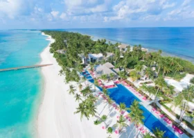 Veduta con drone dell'isola allungata di Kandima con la lunga piscina dai lettini fucsia costeggiata da palme da cocco, la spiaggia di sabbia bianca e il pontile che si protende nella laguna turchese fino all'oceano blu. Kandima Maldives, Atollo di Dhaalu, Maldive.