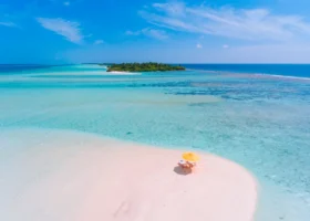 Veduta aerea di una lingua di sabbia bianca isolata con due lettini e un ombrellone giallo, circondata da acque bassissime che sfumano dal turchese chiaro al blu; sullo sfondo un isolotto verde e la barriera corallina. Kandima Maldives, Isola di Kandima, Atollo di Dhaalu, Maldive.