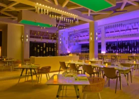 Interno serale di un ristorante di design con tavoli apparecchiati e candele, sedie in rattan e metallo, installazioni di perline sospese dal soffitto e pareti illuminate da luci viola e verdi che creano un'atmosfera vivace e contemporanea. Kandima Maldives, Kandima, Atollo di Dhaalu.
