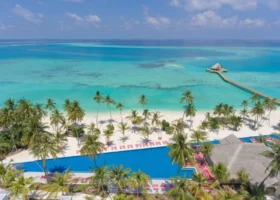 Veduta aerea della spiaggia di sabbia bianca e della lunga piscina bordata di lettini fucsia e palme da cocco, con il padiglione dal tetto in strame e il pontile che raggiunge un gazebo overwater sulla laguna turchese screziata dal reef. Kandima Maldives, Isola di Kandima, Atollo di Dhaalu, Maldive.