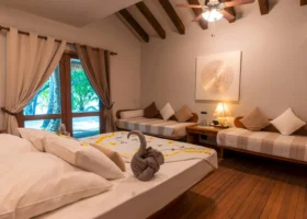 Interno di un Beach Bungalow del NAKAI Alimathà Resort sull'isola di Alimathà, atollo di Vaavu, Maldive: letto matrimoniale con lenzuola bianche decorate da petali gialli di frangipani e un cigno di asciugamani, soffitto in legno con travi a vista e ventilatore a pale, due divani-letto con cuscini a righe, quadro tondo texturizzato e porta-finestra affacciata sul giardino tropicale con palme.