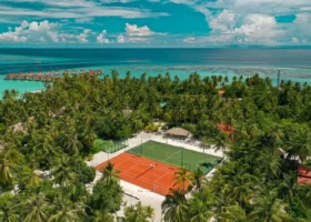 Vista aerea degli impianti sportivi del NAKAI Alimathà Resort sull'isola di Alimathà, atollo di Vaavu, Maldive: campo polivalente da tennis e calcetto con superficie arancione e verde immerso tra le palme da cocco, con sullo sfondo la fila di water villa overwater dal tetto rosso e la laguna turchese che si apre sull'Oceano Indiano.