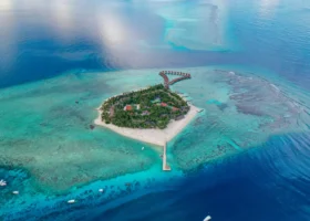 Vista aerea con drone dell'isola di Alimathà, atollo di Vaavu, Maldive, sede del NAKAI Alimathà Resort: la conformazione a forma di cuore coperta di palme, le spiagge di sabbia bianca e il pontile, con la fila curva di water villa overwater che si protende sulla laguna turchese circondata dalla barriera corallina e dall'Oceano Indiano blu intenso.
