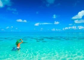 Donna con costume arancione e ampio cappello di paglia verde immersa fino alla vita nella laguna dalle acque cristalline e turchesi di fronte al NAKAI Alimathà Resort, isola di Alimathà, atollo di Vaavu, Maldive, con il fondale sabbioso trasparente, chiazze di corallo affioranti e il cielo azzurro limpido punteggiato di nuvole all'orizzonte dell'Oceano Indiano.